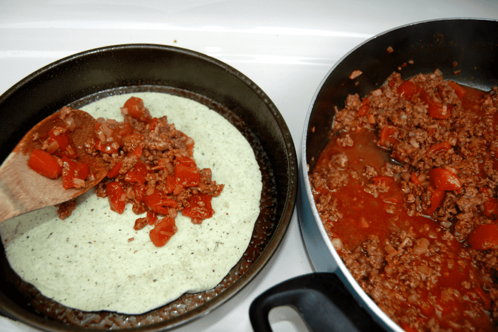 pot of seasoned ground beef beside a pan with the seasoned ground beef getting spooned on top of a tortilla