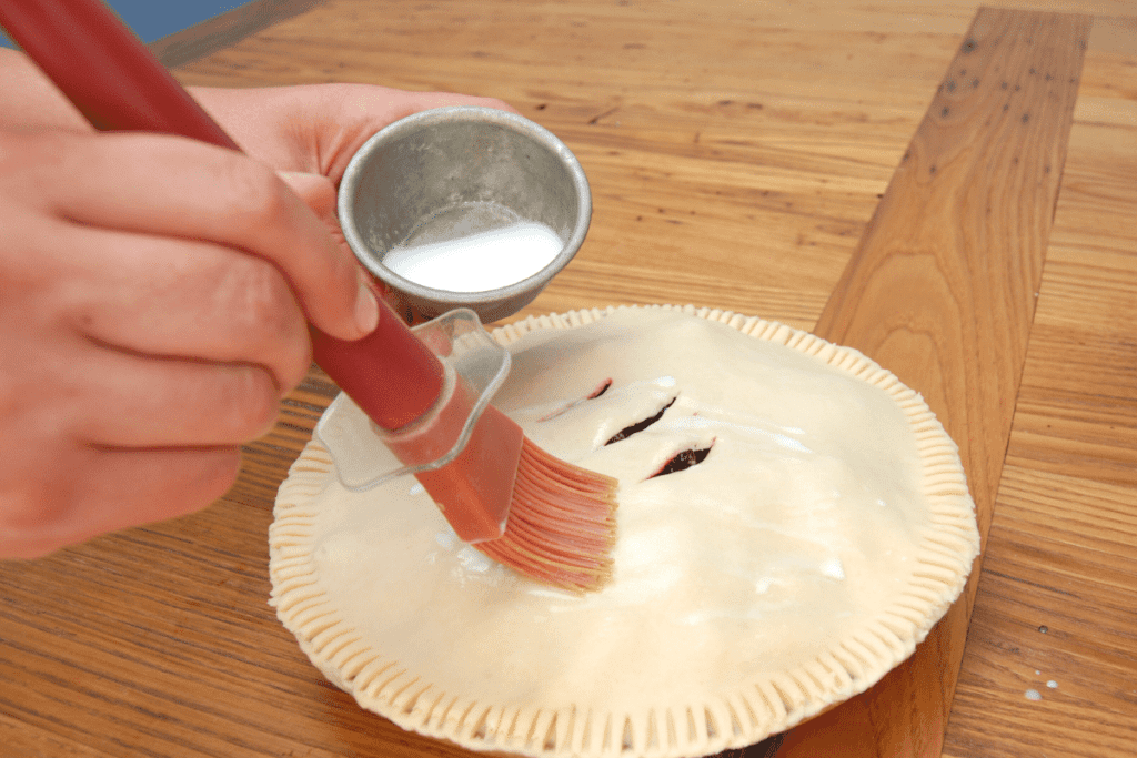 close up of hands using a basting brush to put milk on uncooked pie crust