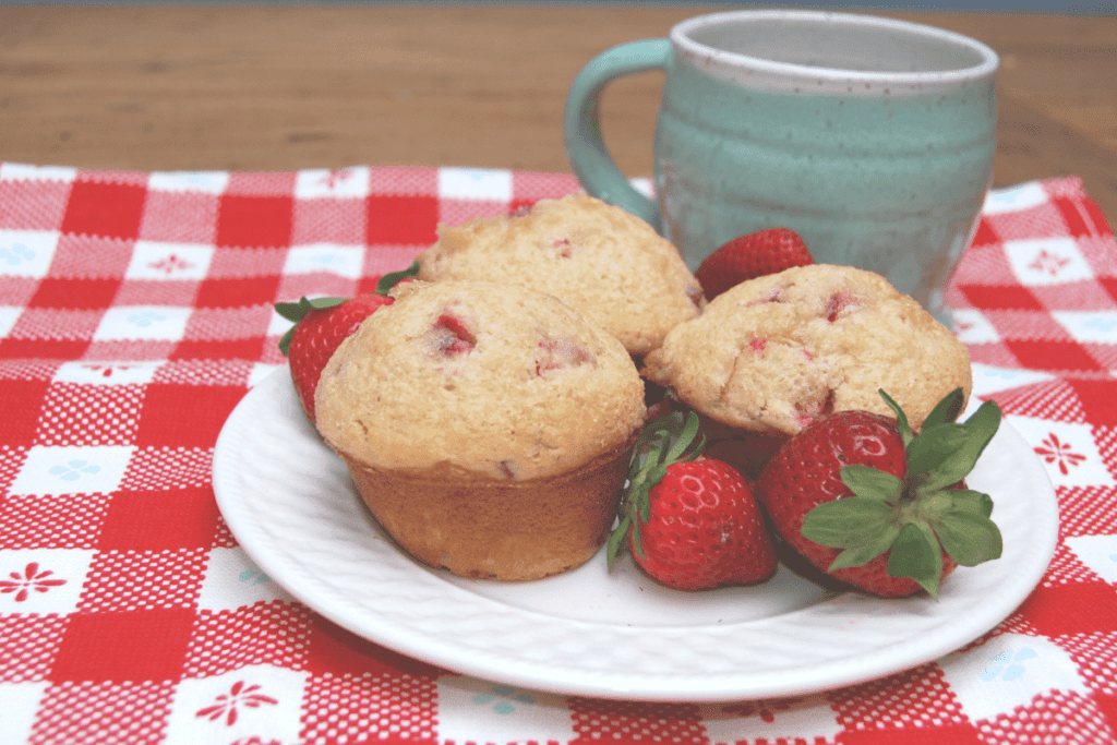 plate with strawberry muffins and strawberries in front of a mug on a checked kitchen towel